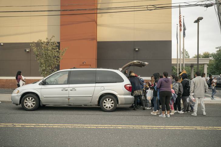 A large huddle of people gather their belongings from the trunk of a gray minivan outside of a hotel in Staten Island.